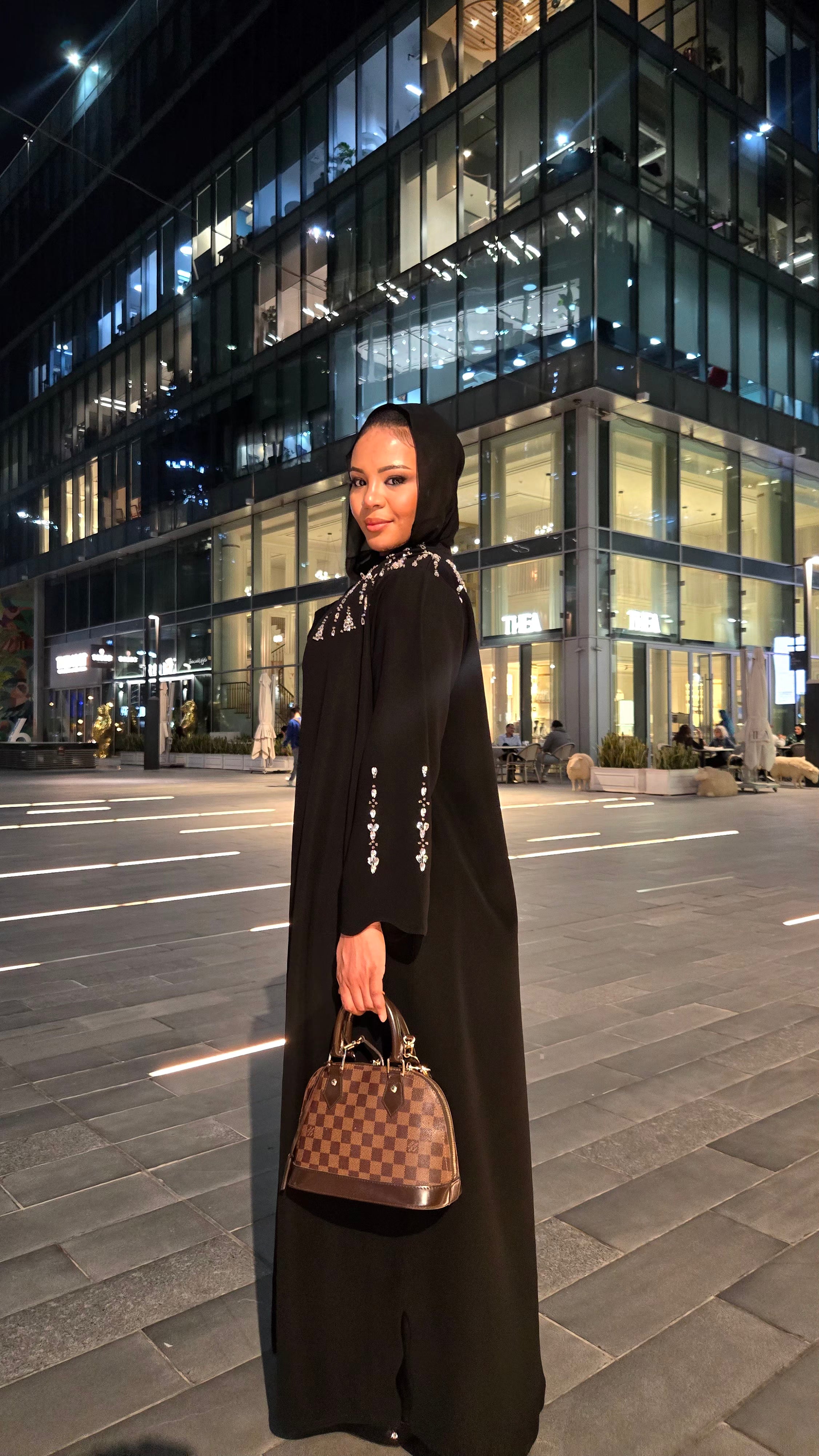 Woman in a black abaya with a patterned scarf holding a handbag, standing on a city street at night.