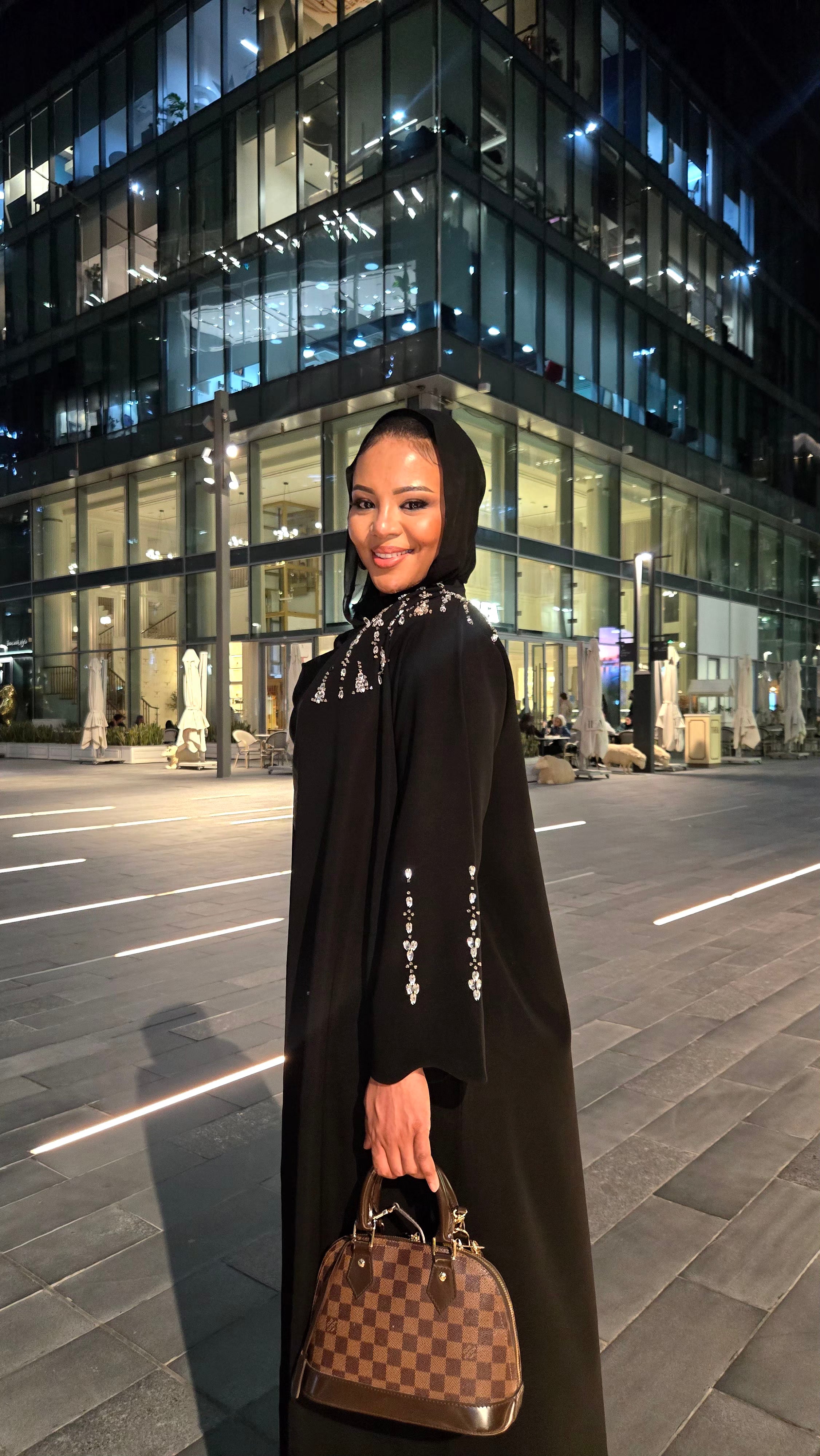 Woman in a long black coat with a patterned scarf and handbag standing on a city street at night.